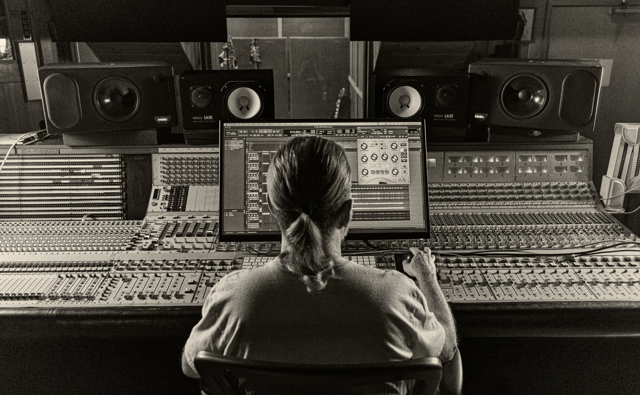 Black and white photo of a man working in front of a large API console with computer monitor on top displaying the Deco Plugin in a DAW.